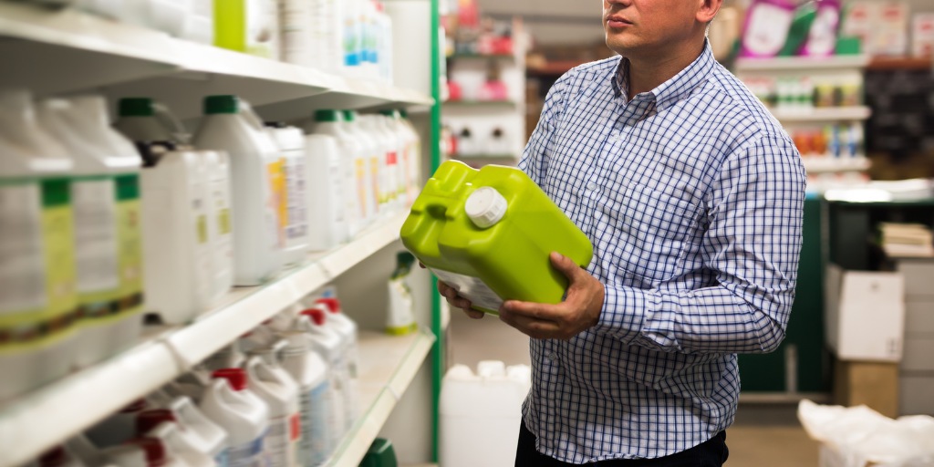 A man in a checkered shirt examines a green jug of liquid fertilizer in a store aisle lined with garden chemical products.