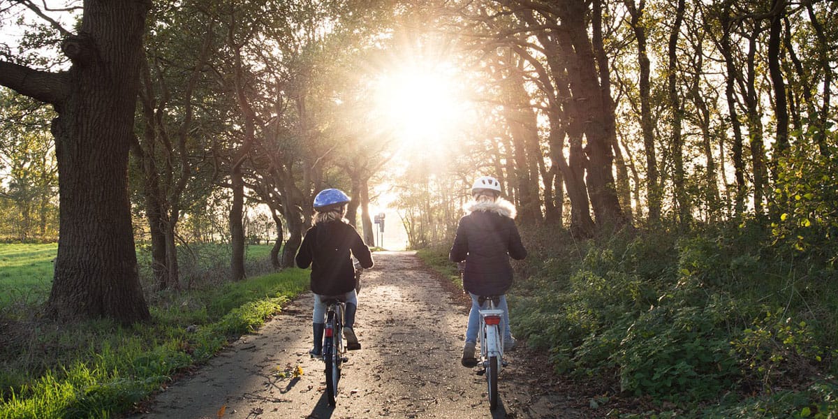 Two young girls wearing helmets bike through a grove of trees towards the setting sun