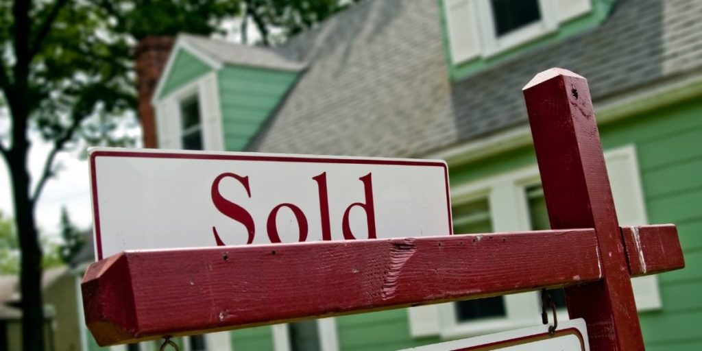 A sign reading "sold" in front of a green house surrounded by trees.